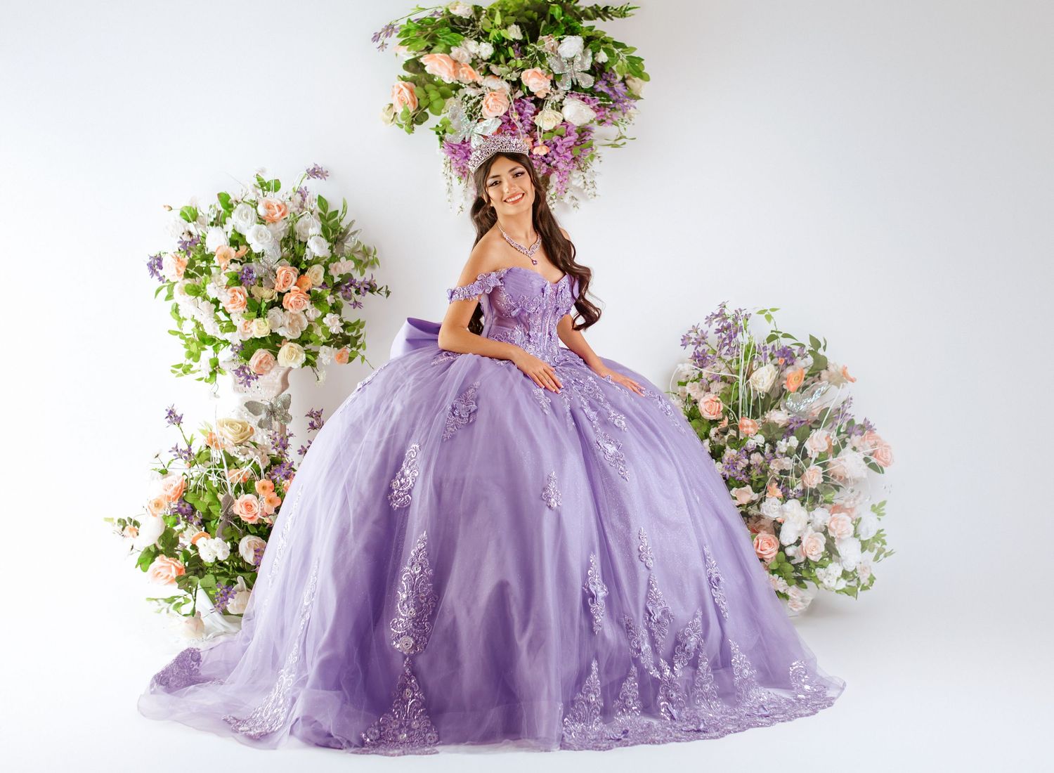 Young woman in lavender ball gown quinceañera dress with floral appliques poses among pink and white flower arrangements.