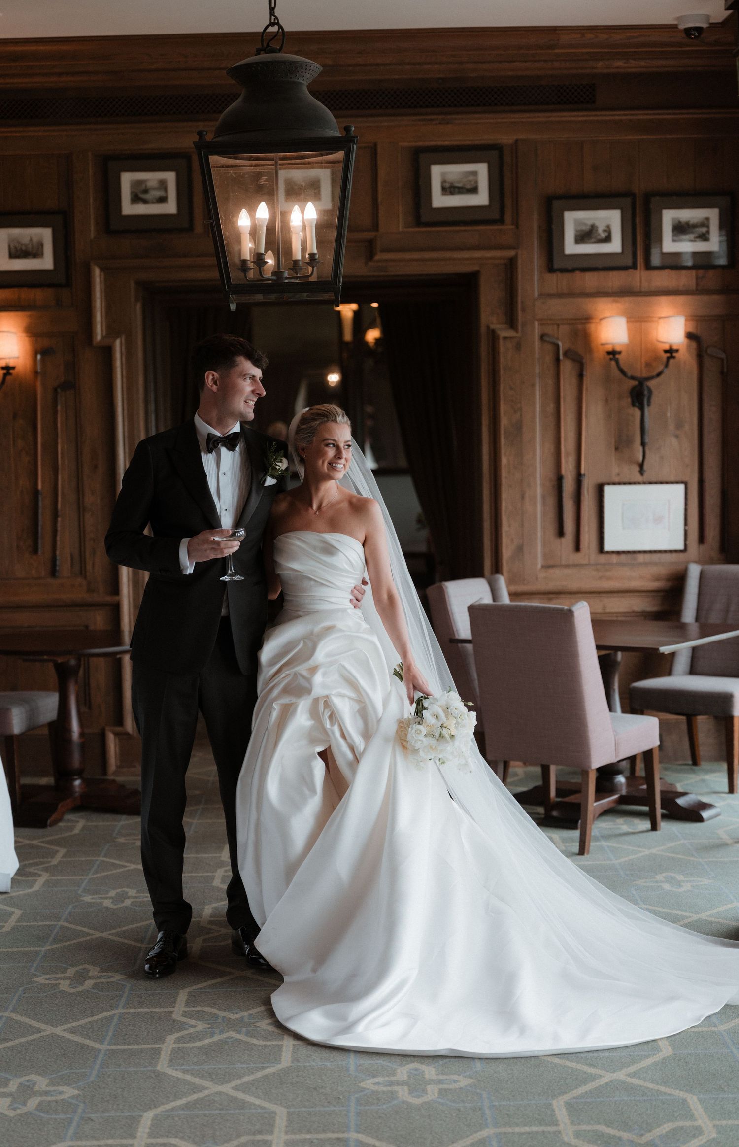 Bride in flowing white gown with long train and groom in tuxedo pose on red carpet steps at elegant wedding venue.