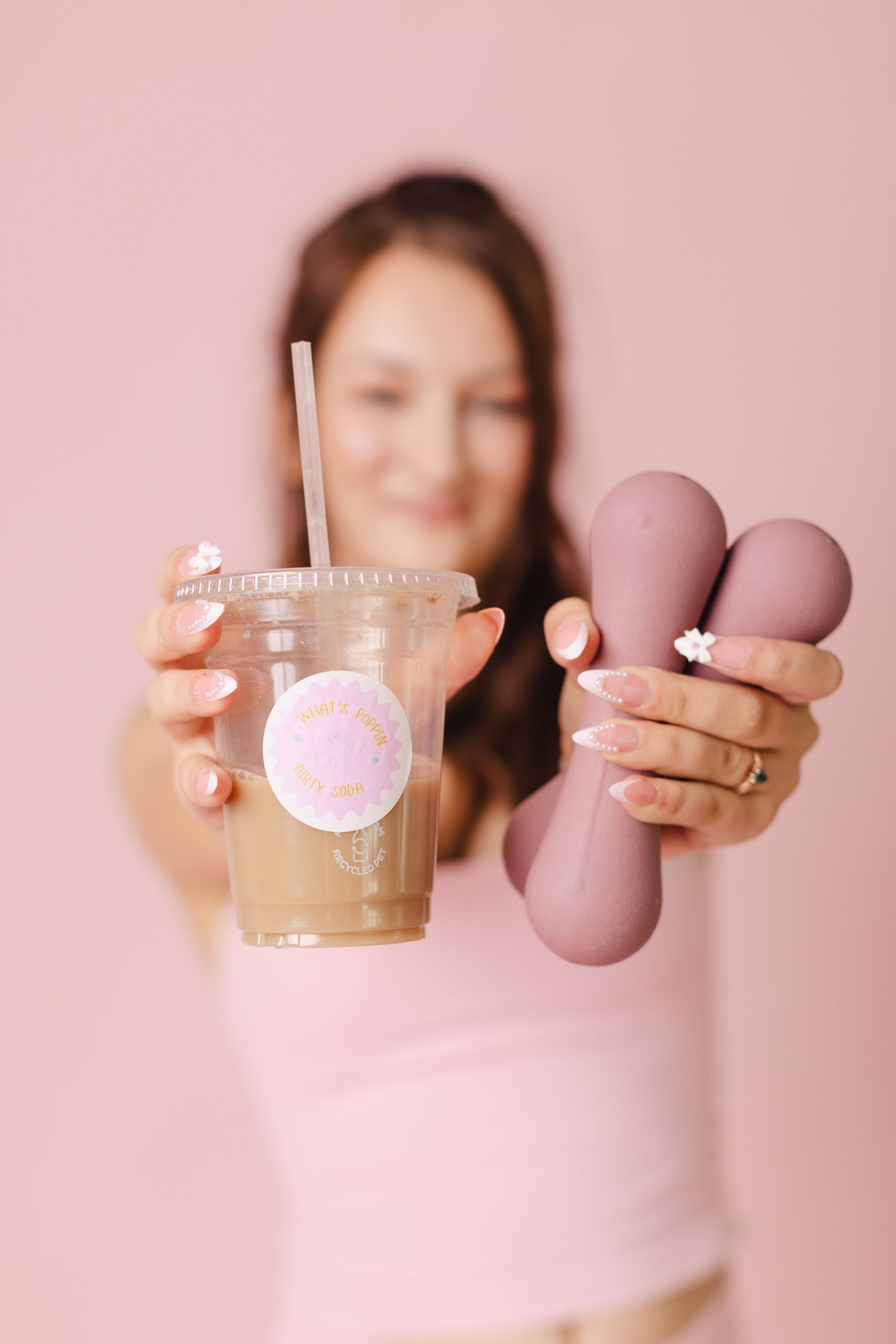 Woman holding iced coffee drink and pink macarons against a soft pink background with manicured nails.