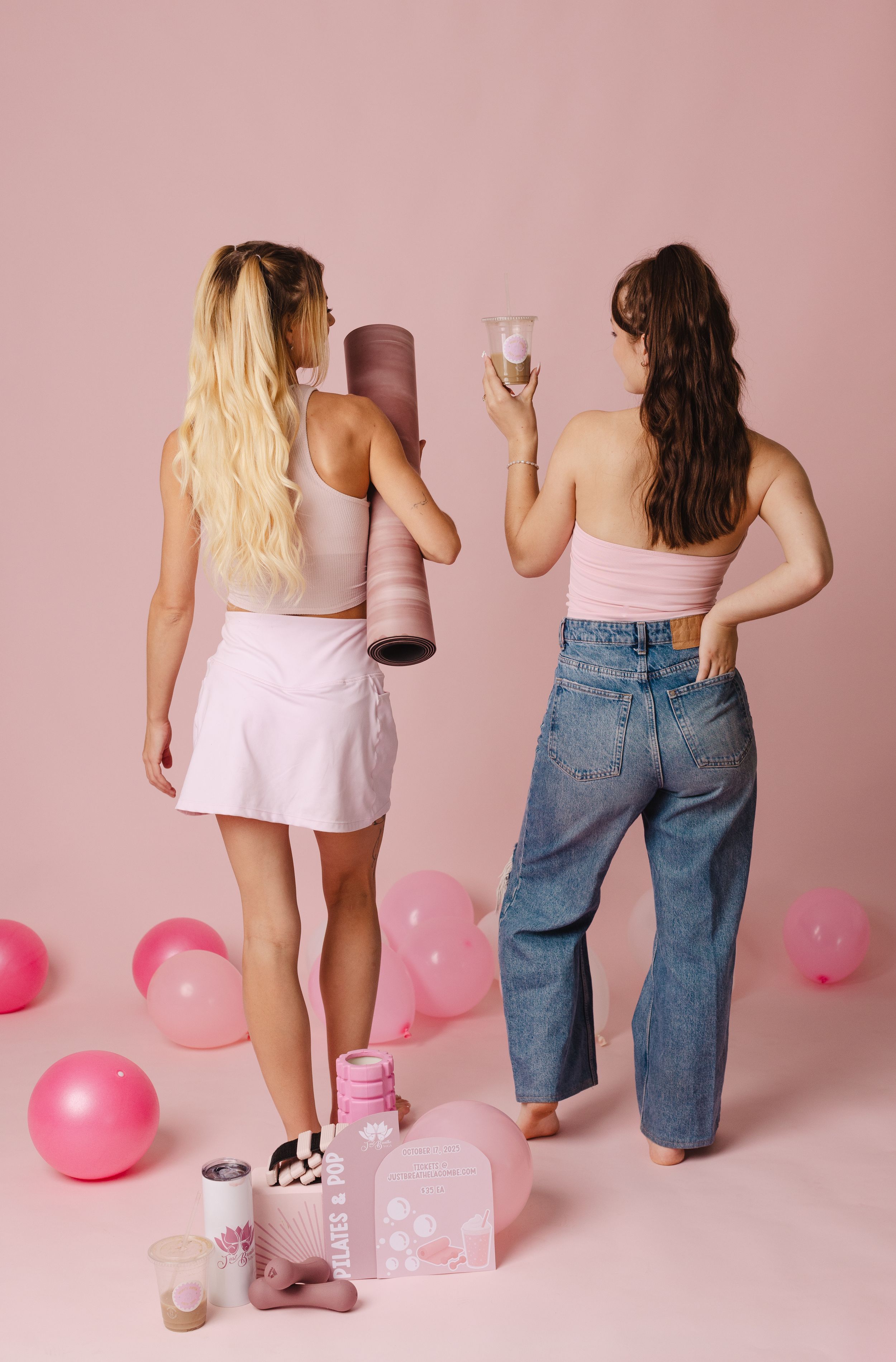 Two women in pink outfits pose with balloons, a yoga mat, and gifts on a pink background.