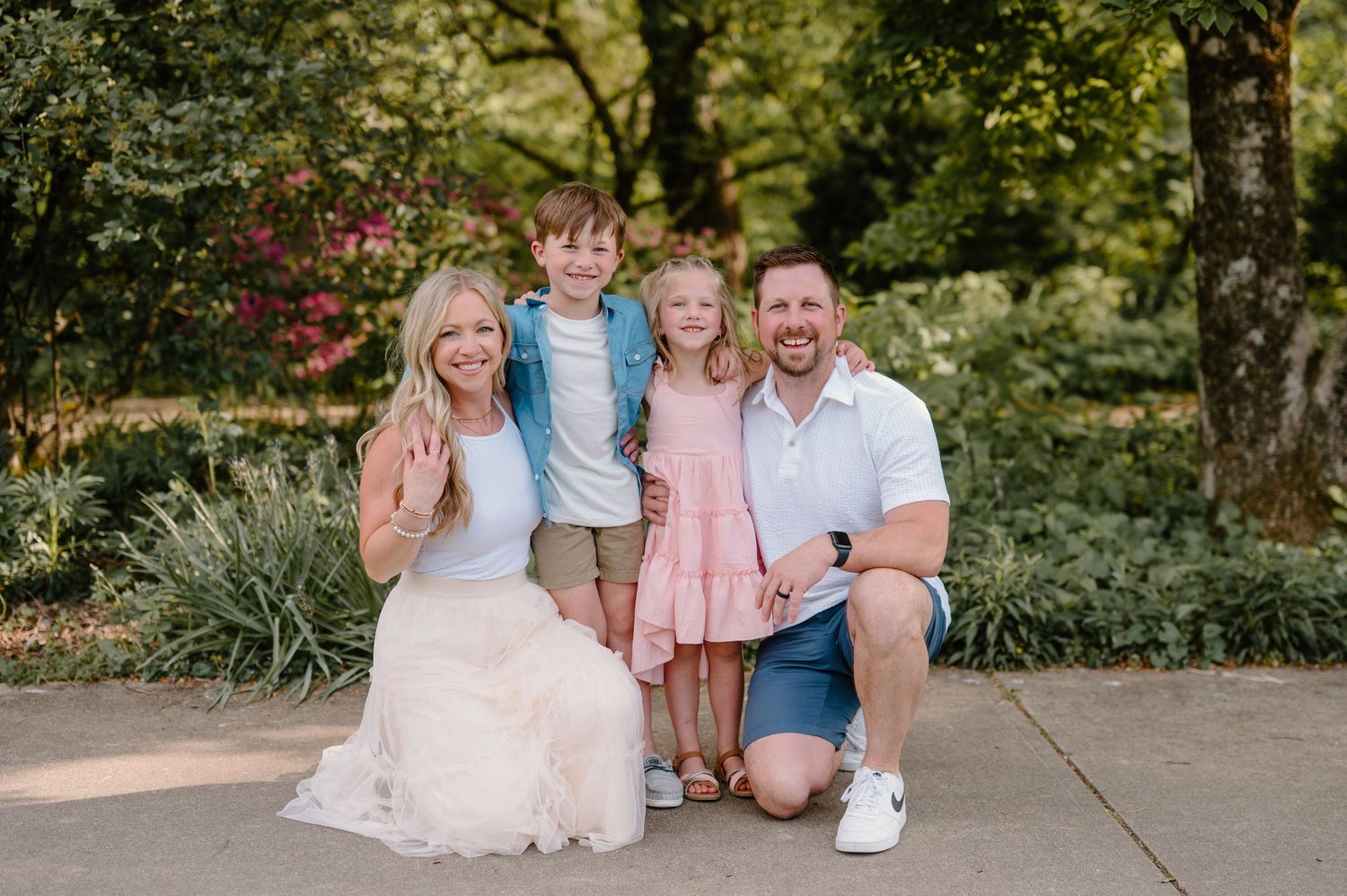 A smiling family of four poses outdoors in a lush garden, with two young children between their parents.