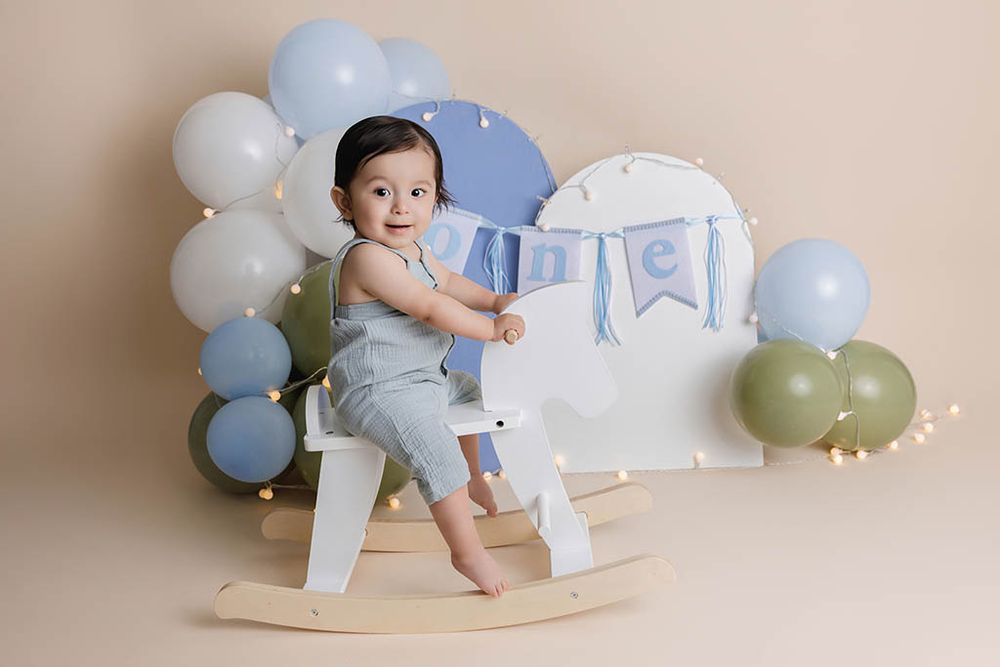 Cute baby boy in gray romper sitting on wooden rocking horse with blue balloon arch backdrop.
