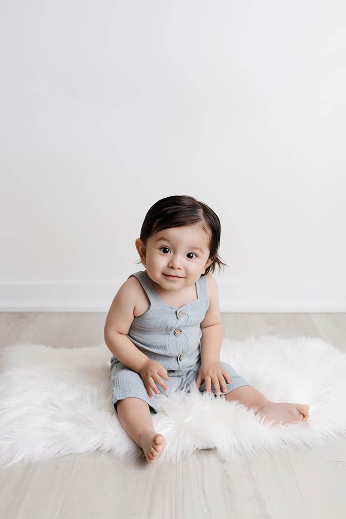 Cute baby in a blue romper sitting on a fluffy white rug smiling at camera, by affordable infant photographer in Menifee.