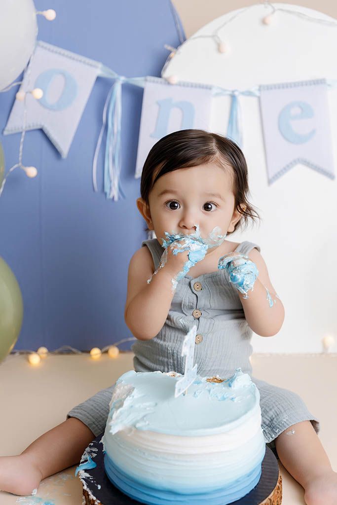 Baby boy in romper smashing blue ombre first birthday cake during one year cake smash photo session, by affordable photographer in Menifee.