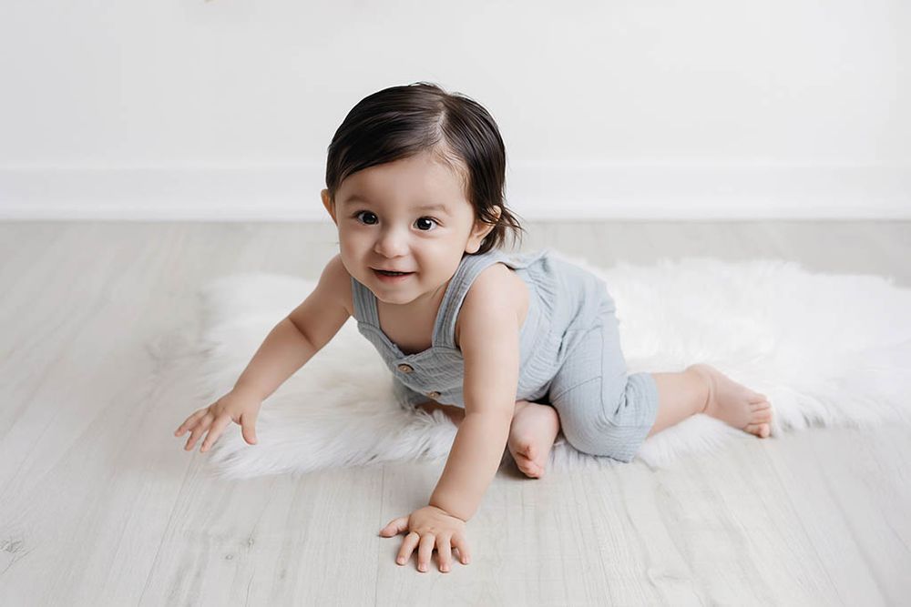A smiling baby in a gray romper crawls on a white fluffy rug over light hardwood floors in a bright room.