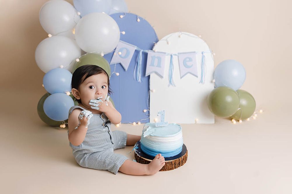 Baby boy in gray romper smiles during first birthday cake smash with blue balloons and 'one' banner.