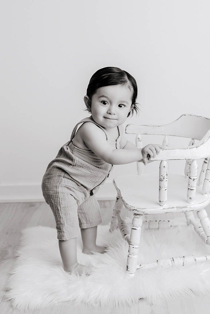 Black and white photo of a smiling baby standing beside a white vintage chair on a fluffy rug.
