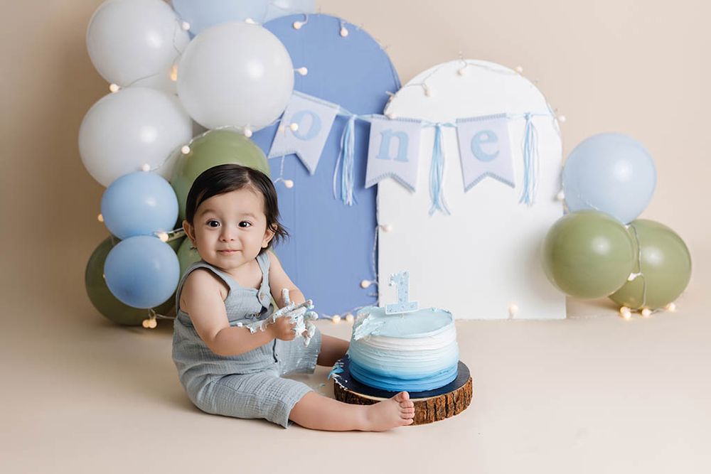 Baby boy in gray romper smiles at cake smash session with blue balloons and ONE banner on first birthday.