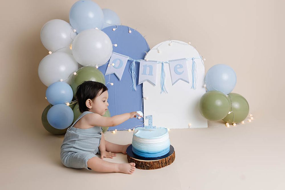 Baby boy in blue romper touches his first birthday cake surrounded by balloon decorations by affordable Menifee photographer.