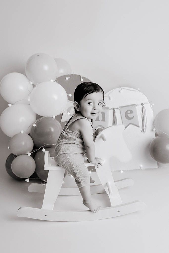 Black and white photo of baby sitting on rocking horse surrounded by balloons at a 1st birthday celebration, by affordable CA photographer.