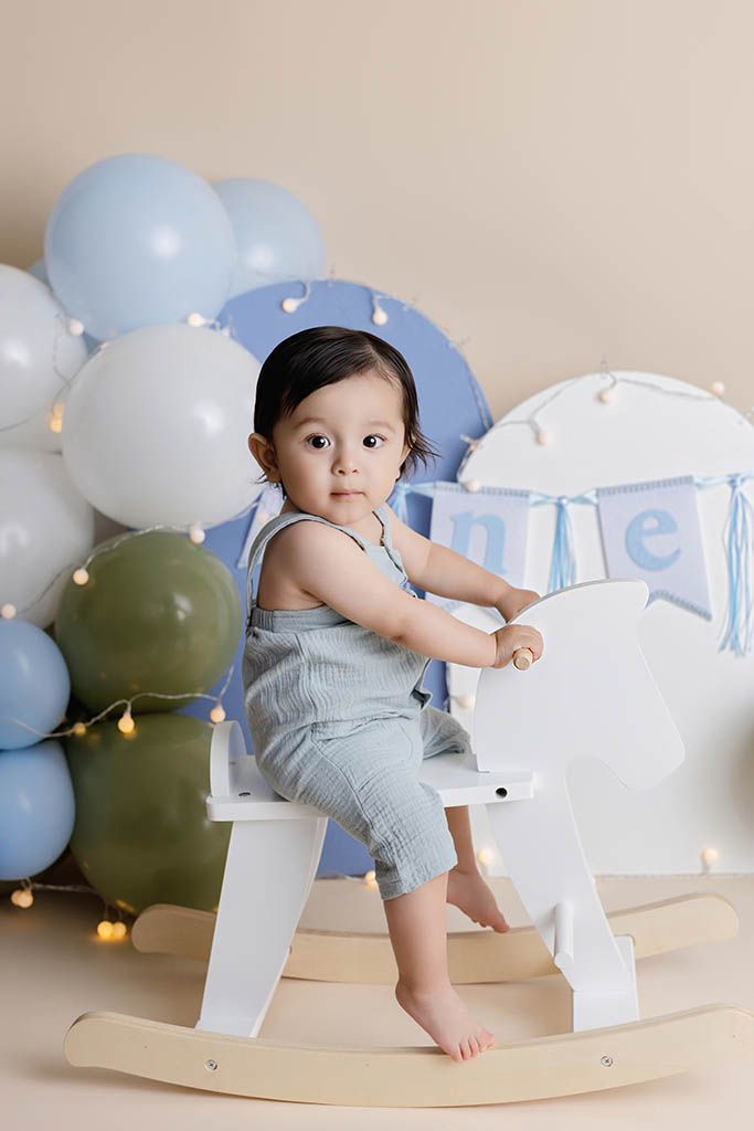 Baby boy in blue romper sits on white rocking horse surrounded by blue and white balloons for first birthday.