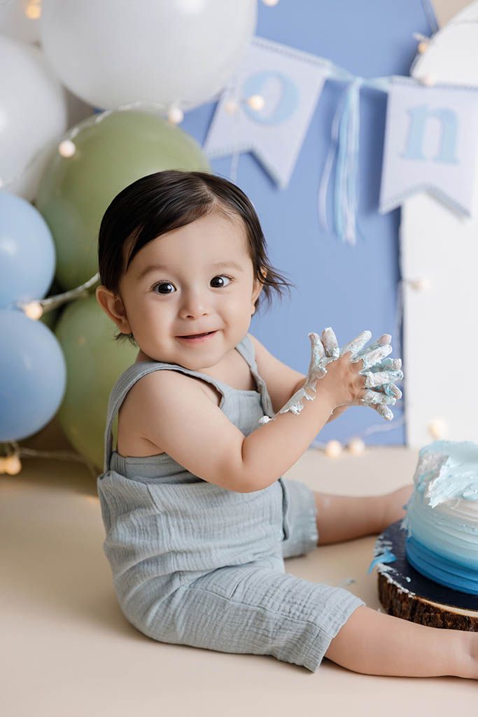 Smiling baby boy in blue romper with frosting on hands beside first birthday cake and balloons.