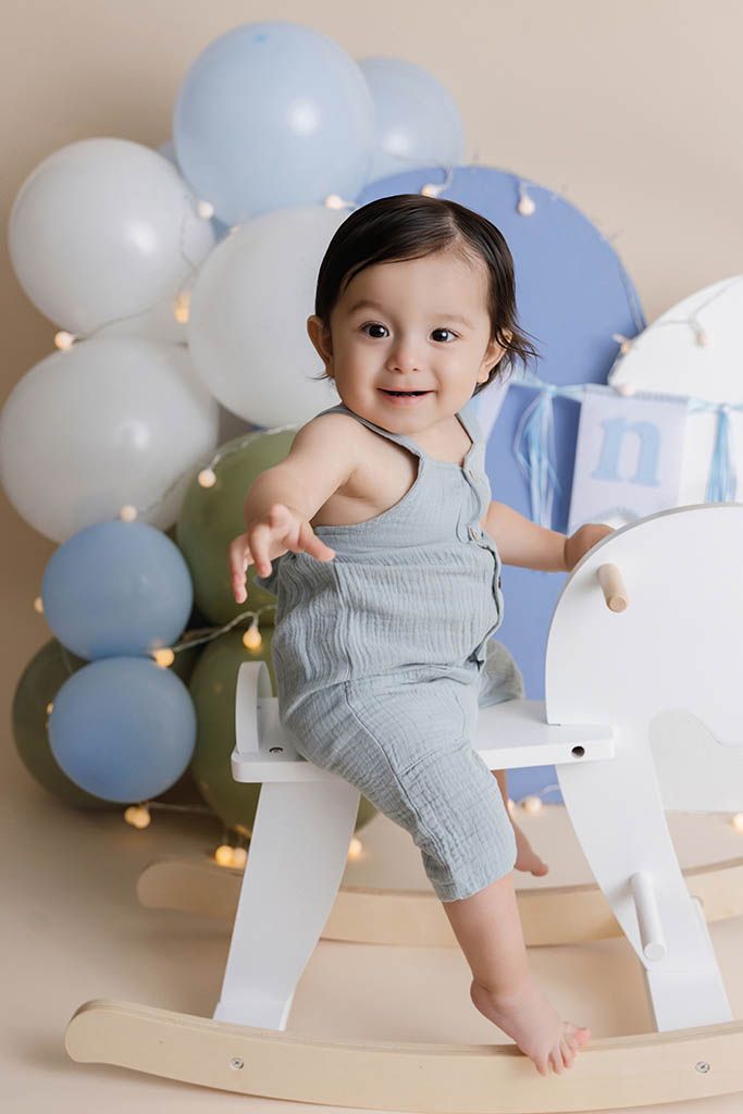 Adorable baby boy in blue linen romper sits on white rocking horse surrounded by balloons, by affordable Menifee photographer .