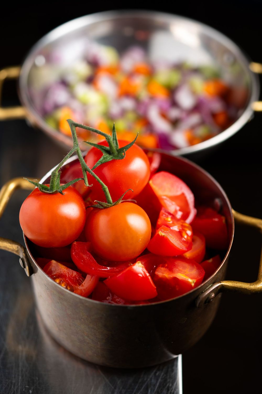 Fresh cherry tomatoes on vine in a copper pot with chopped vegetables in a steel bowl on dark background.