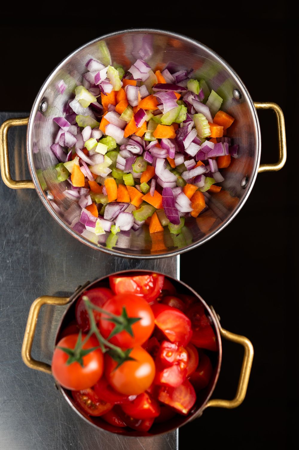 Two silver pots with gold handles filled with fresh chopped vegetables and cherry tomatoes ready for cooking.