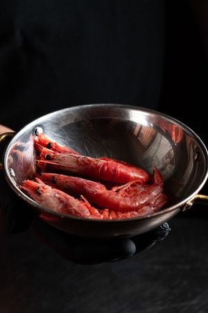 Fresh red prawns displayed in a shiny stainless steel pan against a dark background.