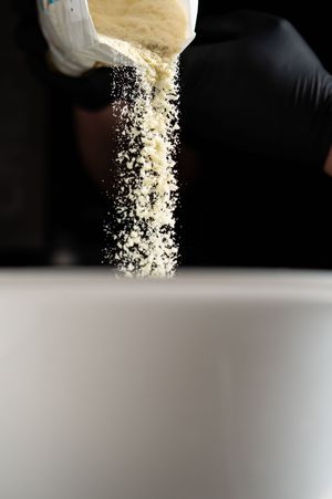 A gloved hand pouring white powder flour into a white bowl against a dark background.