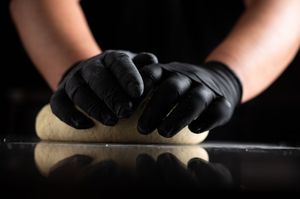 Hands in black gloves kneading dough on a reflective surface against a dark background.