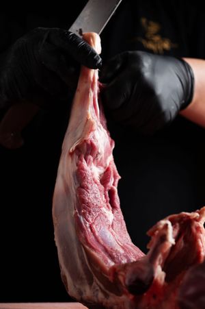 Chef in black gloves cutting raw lamb leg with a sharp knife on dark background.