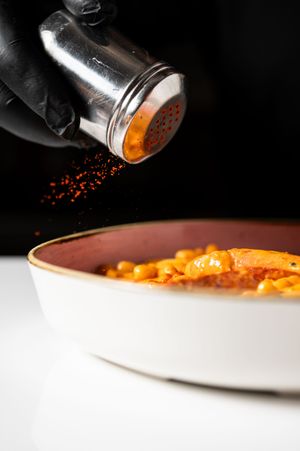 A chef in black gloves sprinkling red spice from a shaker onto a bowl of creamy orange pasta dish.