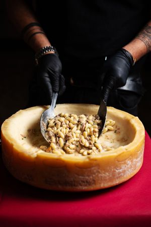 Chef in black gloves mixing pasta inside a hollowed Parmesan cheese wheel on a red tablecloth.