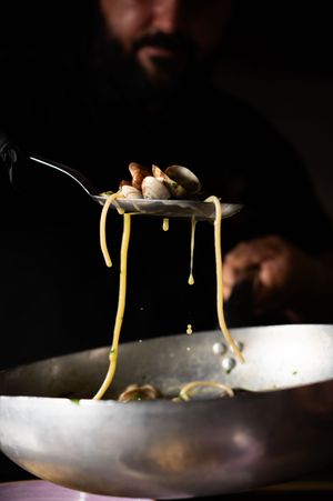Chef lifting spaghetti alle vongole with clams on a fork over a steel pan, sauce dripping dramatically.