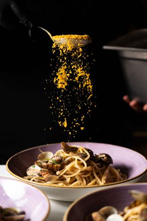 Chef sprinkling golden breadcrumbs over linguine with clams in a purple bowl on dark background.