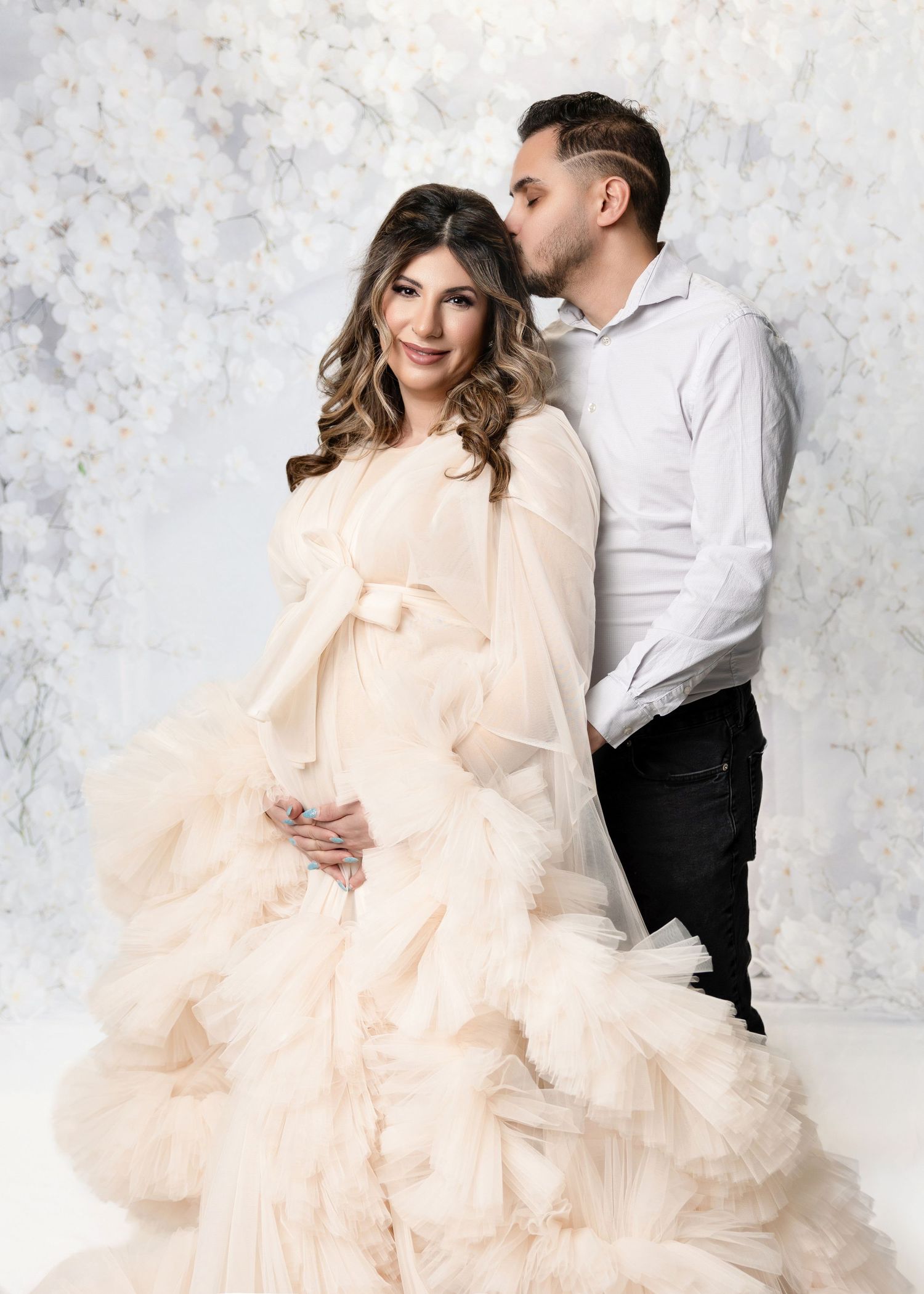Couple in maternity photoshoot, woman in white gown, floral backdrop, tender moment.