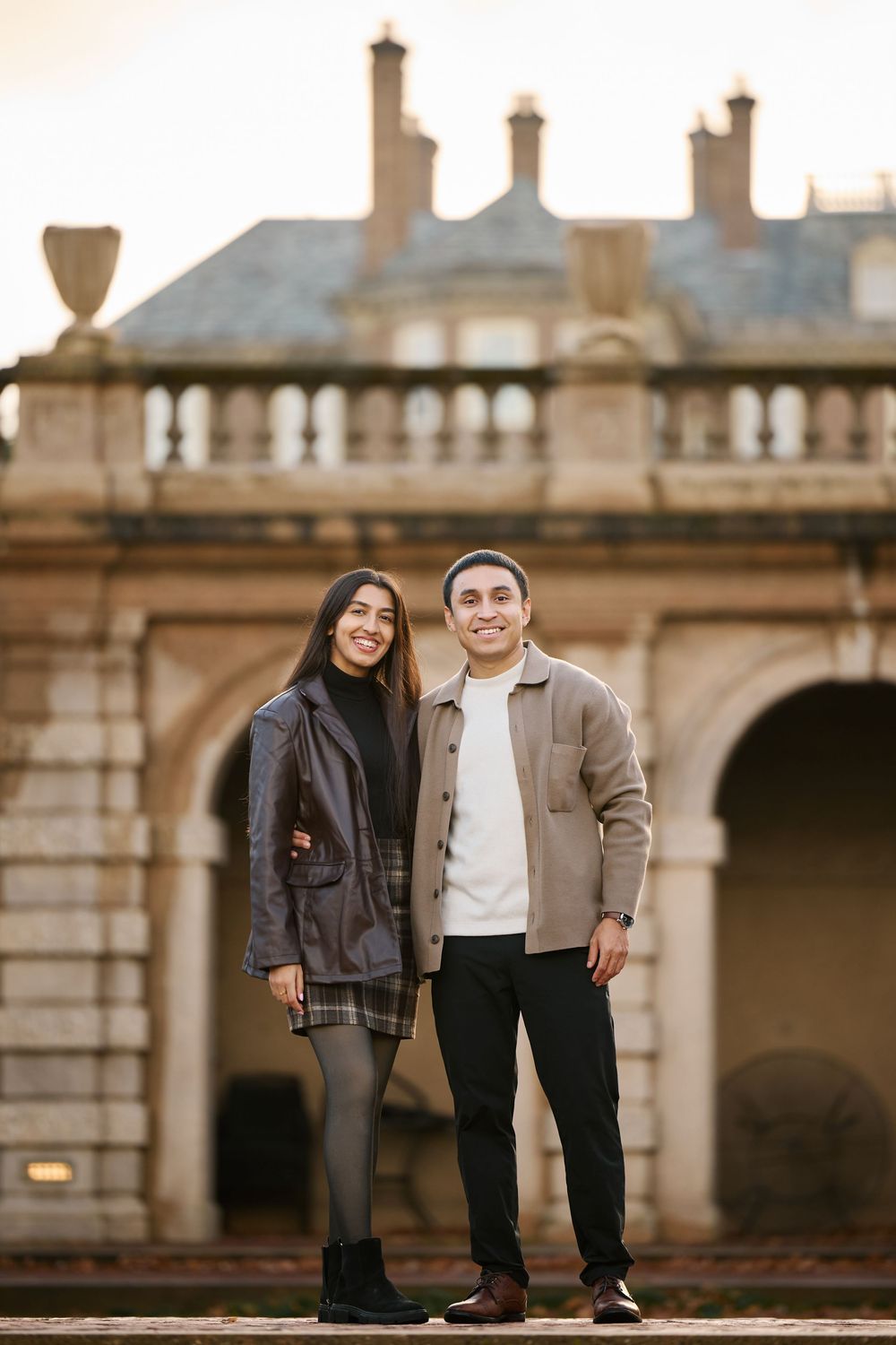Smiling engaged couple posing together in front of historic stone building at golden hour.