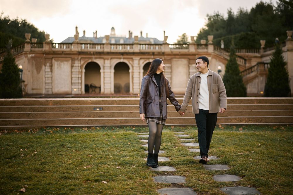 Couple holding hands while walking on garden path with grand historic estate in background.