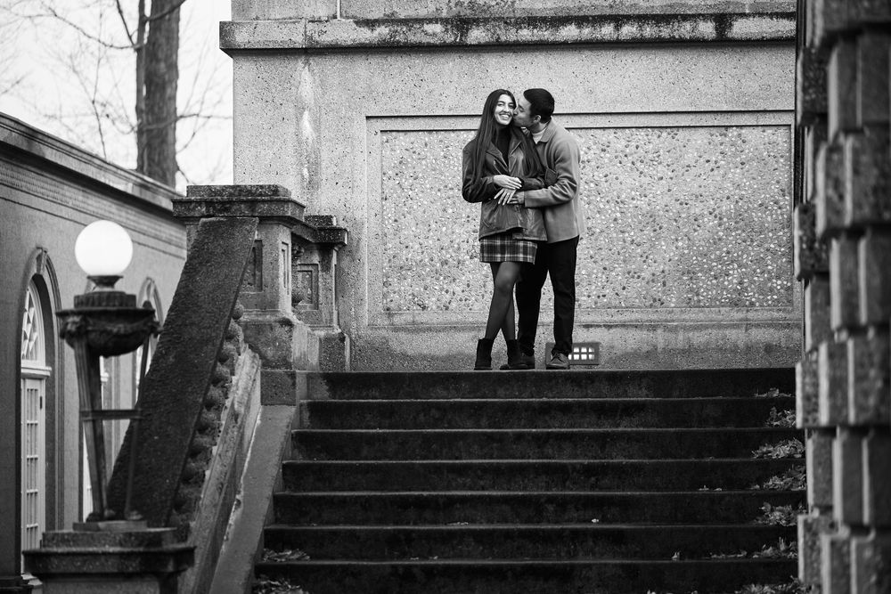 Black and white photo of a romantic couple embracing on stone steps near a classical building.