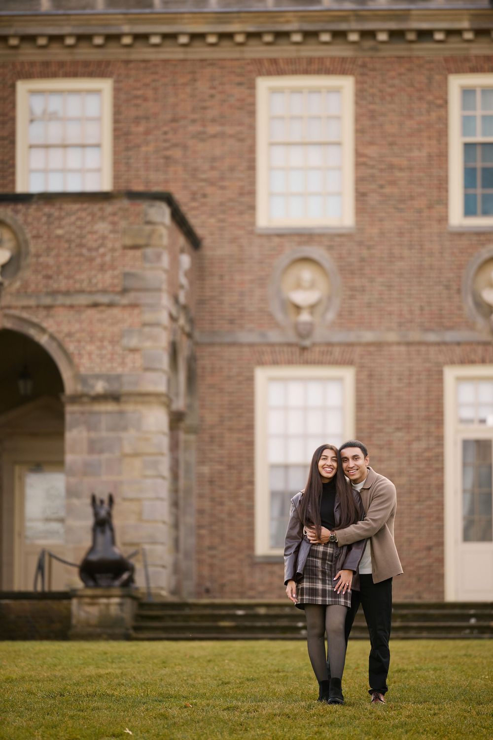 Smiling couple embracing on lawn before classic red brick colonial building.