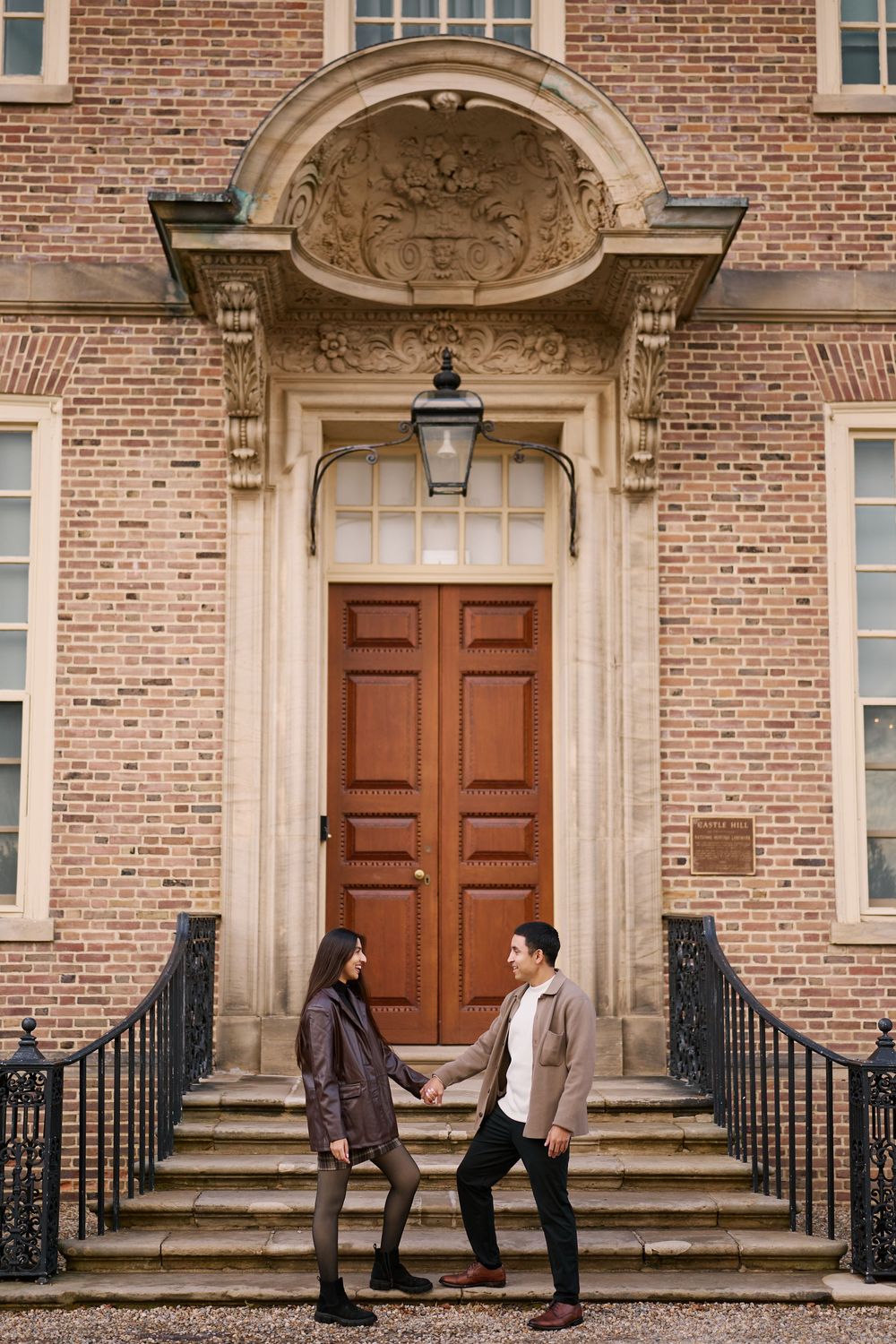 Couple holding hands on steps of elegant brick building with ornate wooden double doors.
