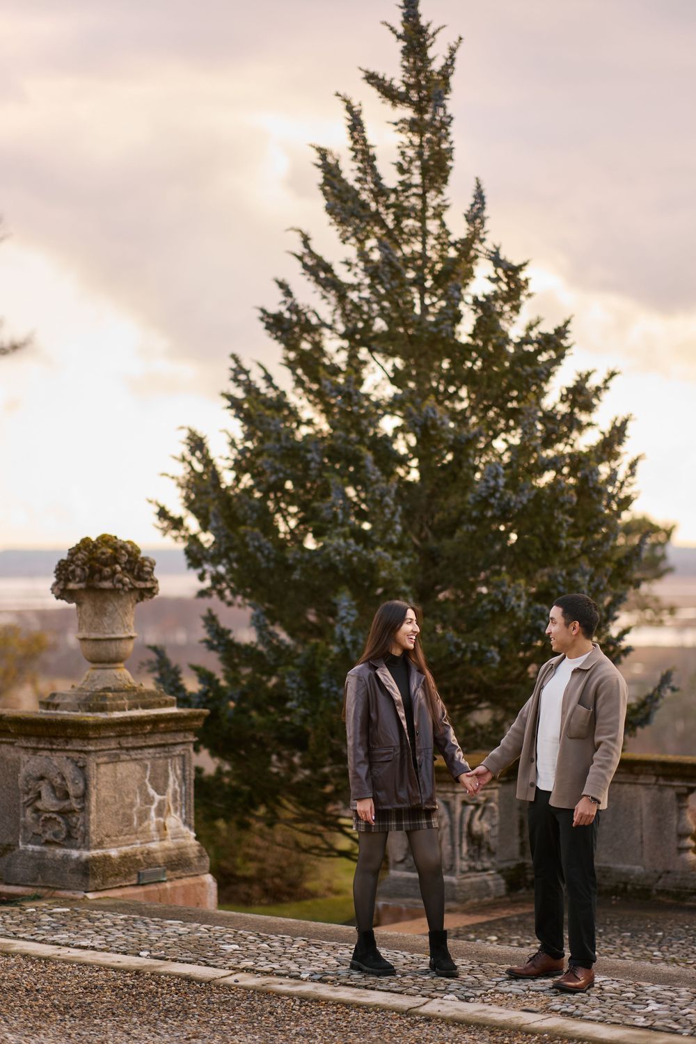 Couple holding hands on a stone terrace with a tall evergreen tree at sunset.