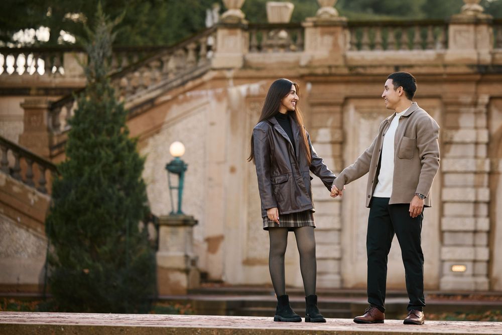 Couple holding hands and smiling at each other outdoors near ornate stone steps and architecture.