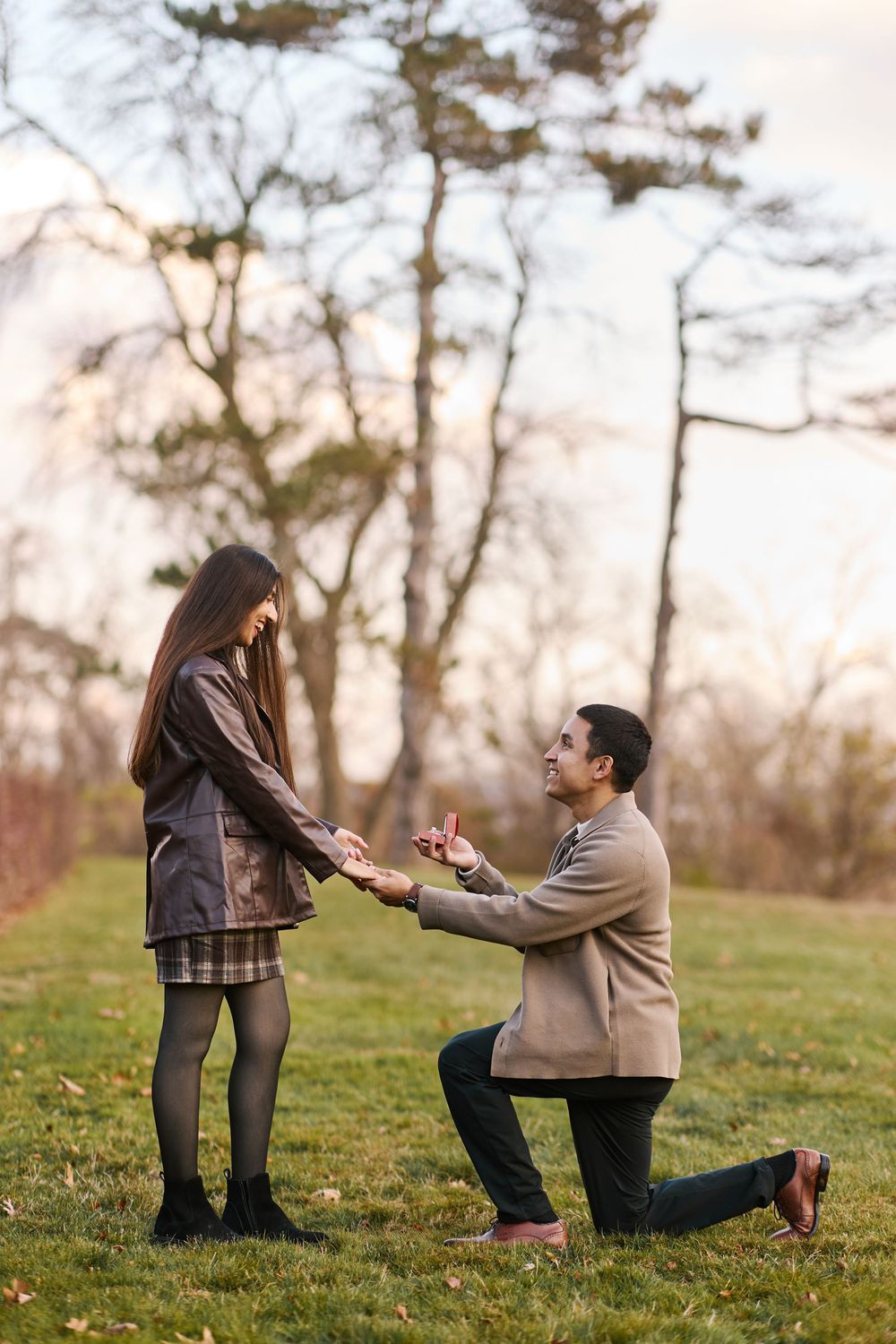 Man proposes on one knee to woman in autumn park, holding ring box while she smiles, surrounded by bare trees.