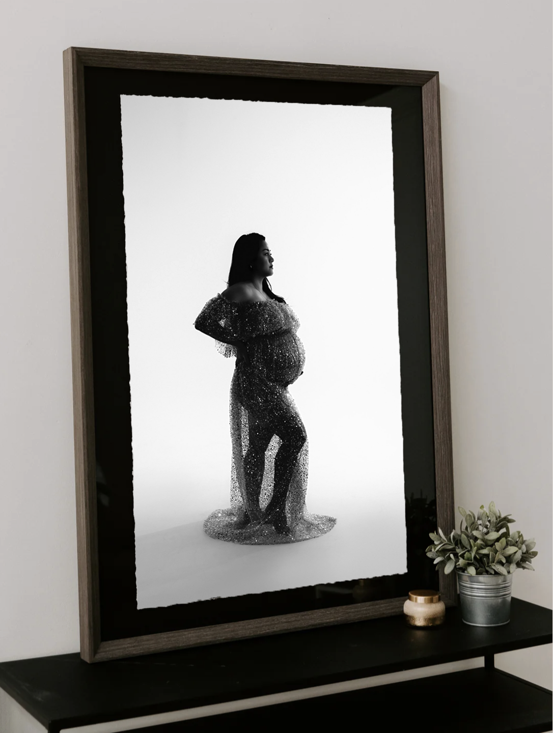 Black and white maternity photo mounted on black in dark wood frame showing pregnant woman in sheer gown, displayed on a side table.
