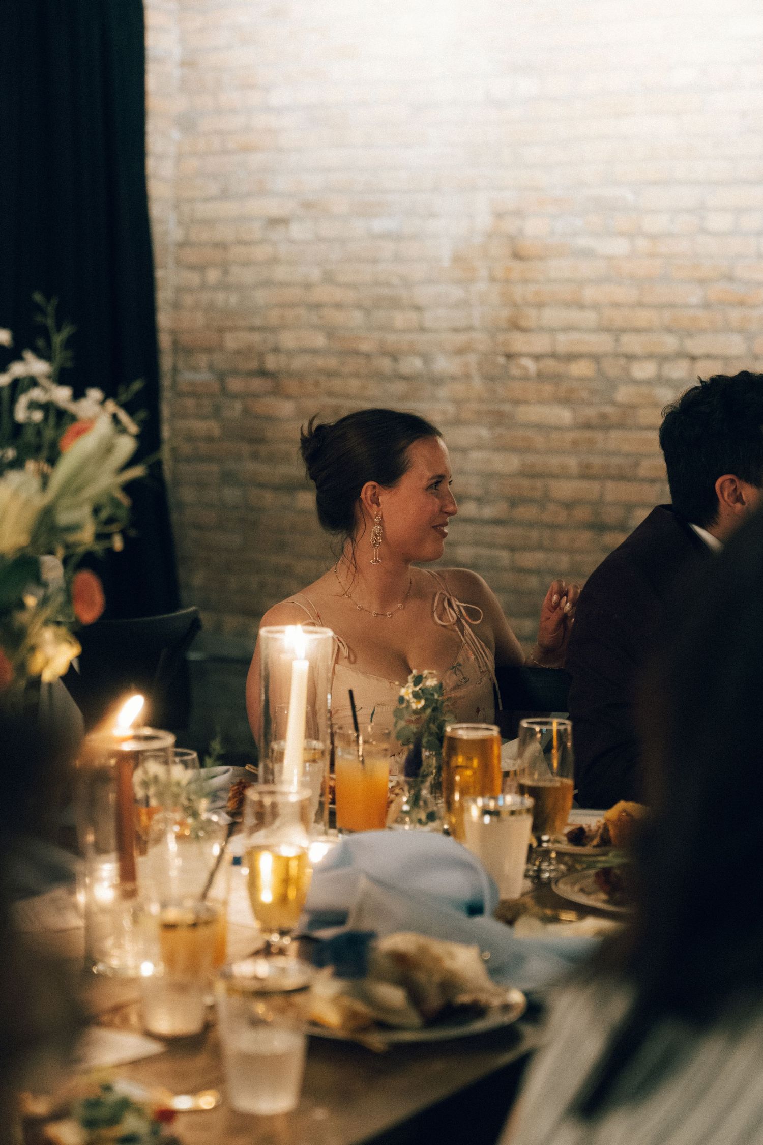 Bride in off-shoulder gown sits at candlelit wedding reception table with guests, glasses, and floral arrangements.