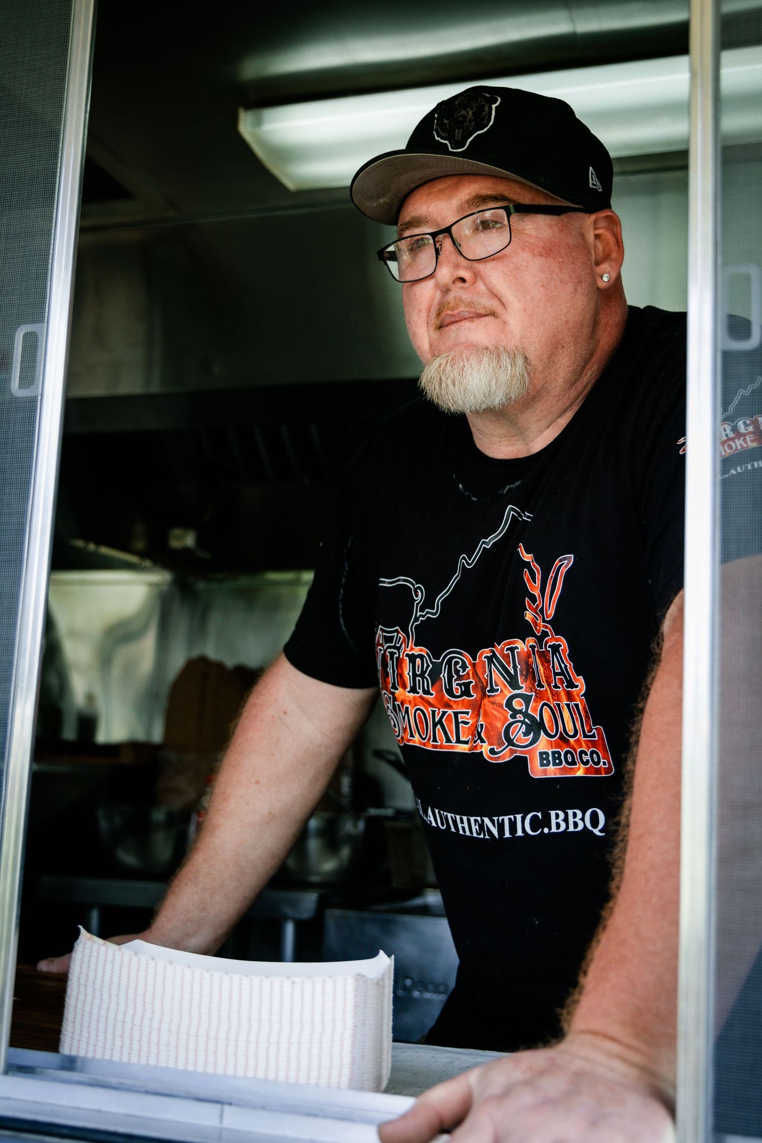 Man in Virginia Smoke & Soul BBQ Co. shirt and cap serves food from a BBQ food truck window.