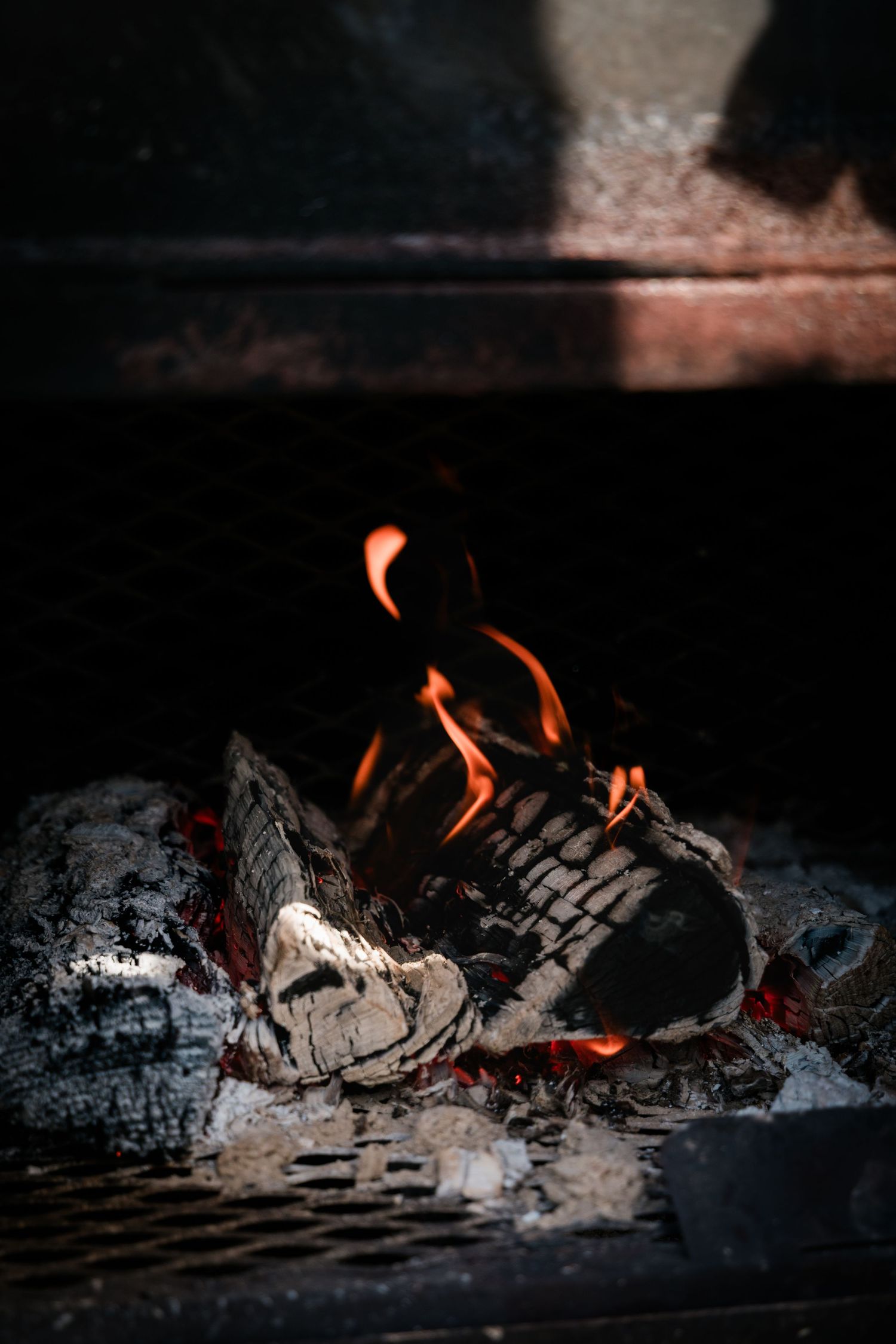 Burning wood logs with orange flames and white ash in a dark fireplace grill.