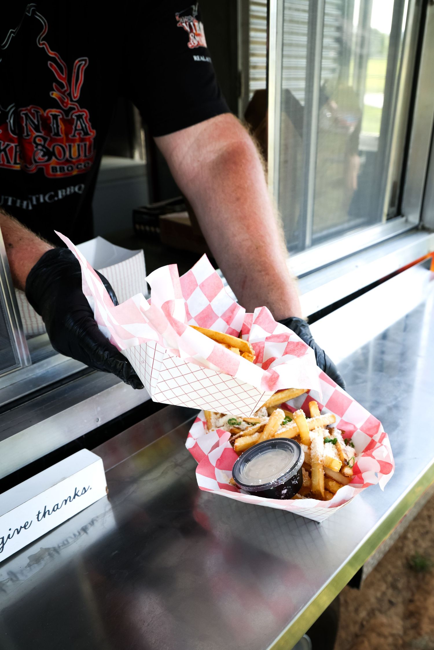 Food truck worker in black gloves serving loaded fries in red checkered paper baskets at a stainless steel counter.