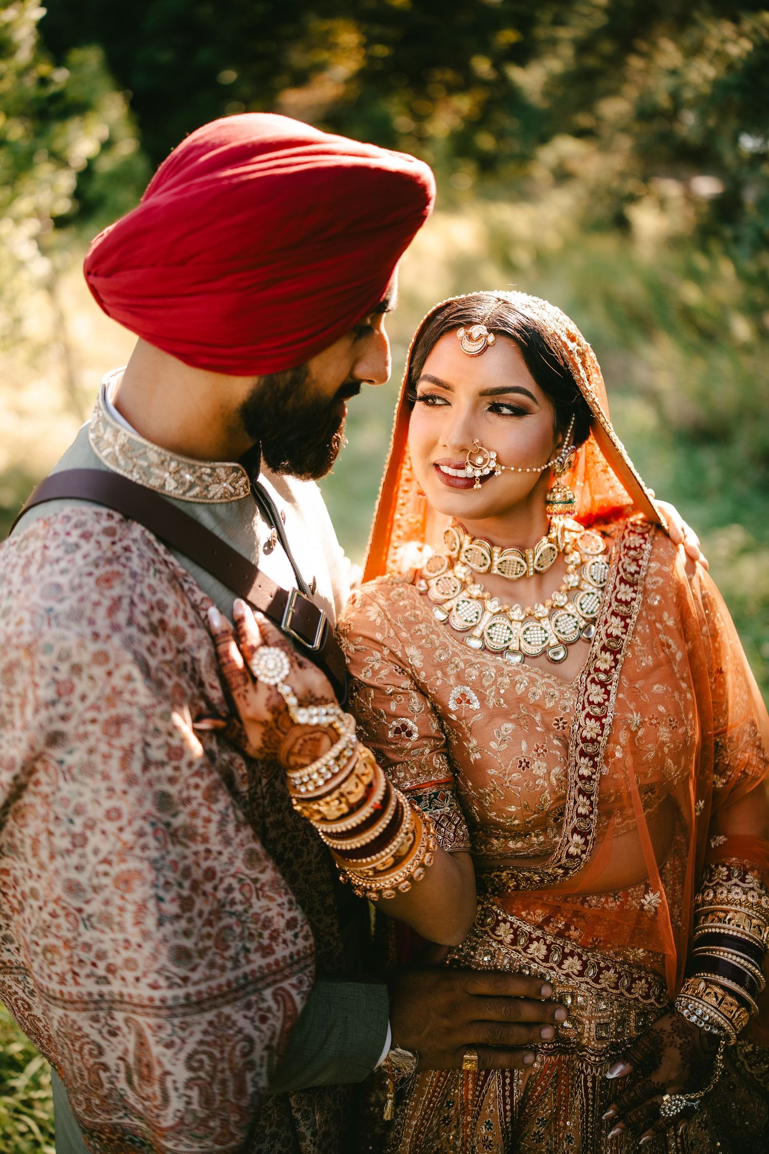 A Sikh bride and groom in traditional attire, she in orange lehenga with gold jewelry, he in red turban, share a loving gaze.