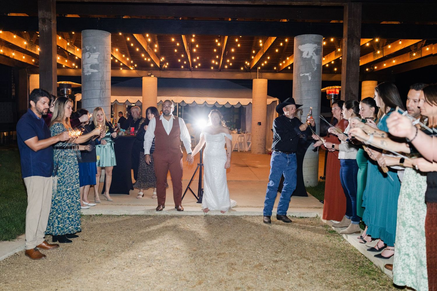 Newlywed couple walks through cheering guests at outdoor wedding reception with string lights and columns.