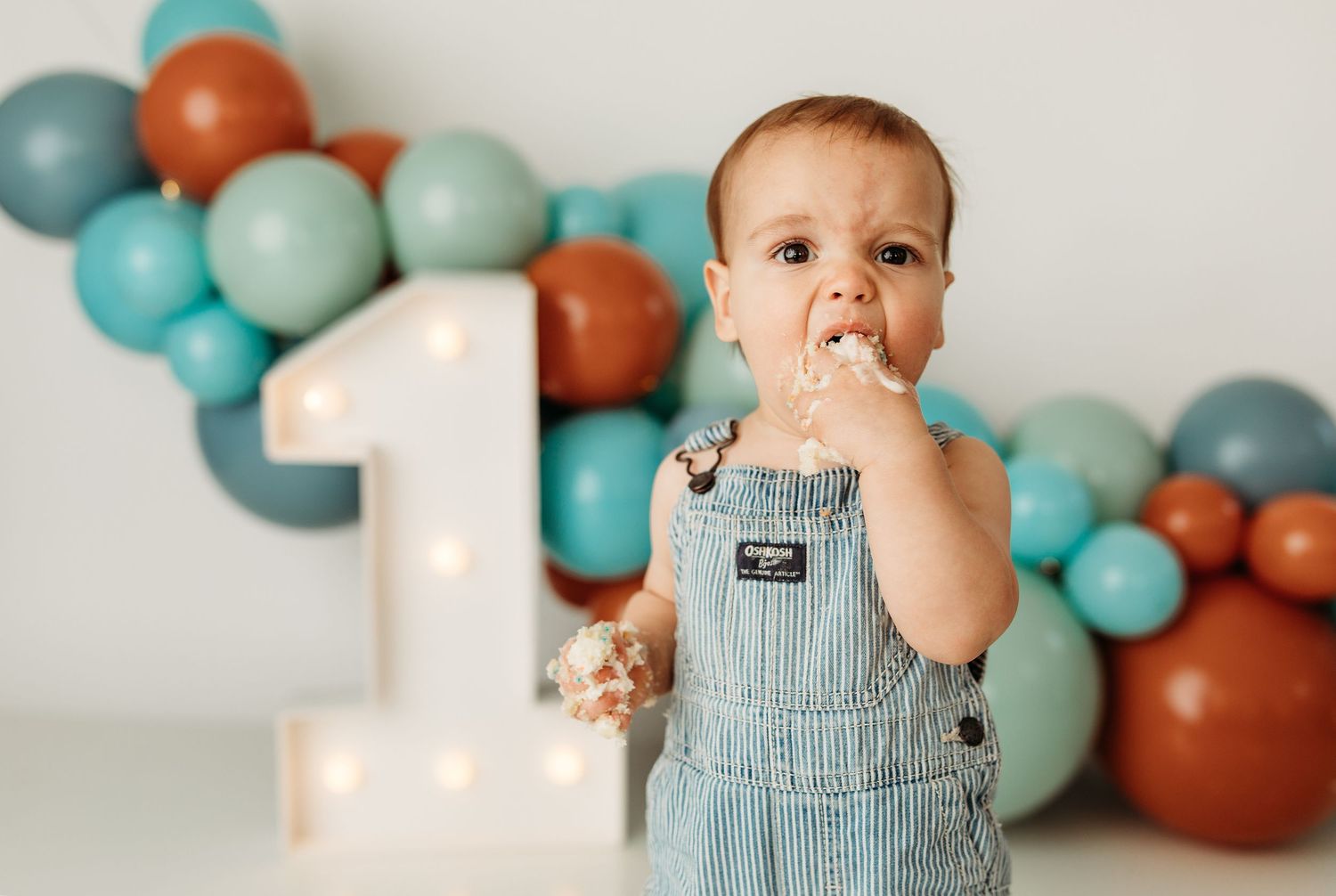 Baby boy in denim overalls celebrates first birthday with colorful balloon garland and illuminated number one backdrop.