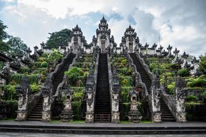 Ancient Balinese Hindu temple with ornate stone carvings, tiered staircases, and lush tropical greenery under cloudy skies.