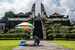 Two people playing a board game under a colorful umbrella in front of a traditional Balinese temple gate.