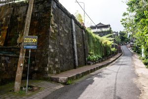 A narrow road runs alongside a moss-covered stone wall with lush greenery and a directional sign in Bali, Indonesia.