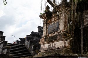Ancient Balinese Hindu temple with ornate stone carvings, moss-covered walls, and hanging vines under a cloudy sky.