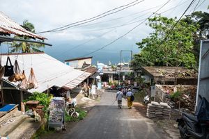 A narrow village street in Bali lined with local shops, power lines overhead, and lush greenery with ocean views.