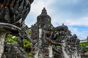 Intricate dark stone Balinese temple sculptures with mythological figures against a cloudy sky.